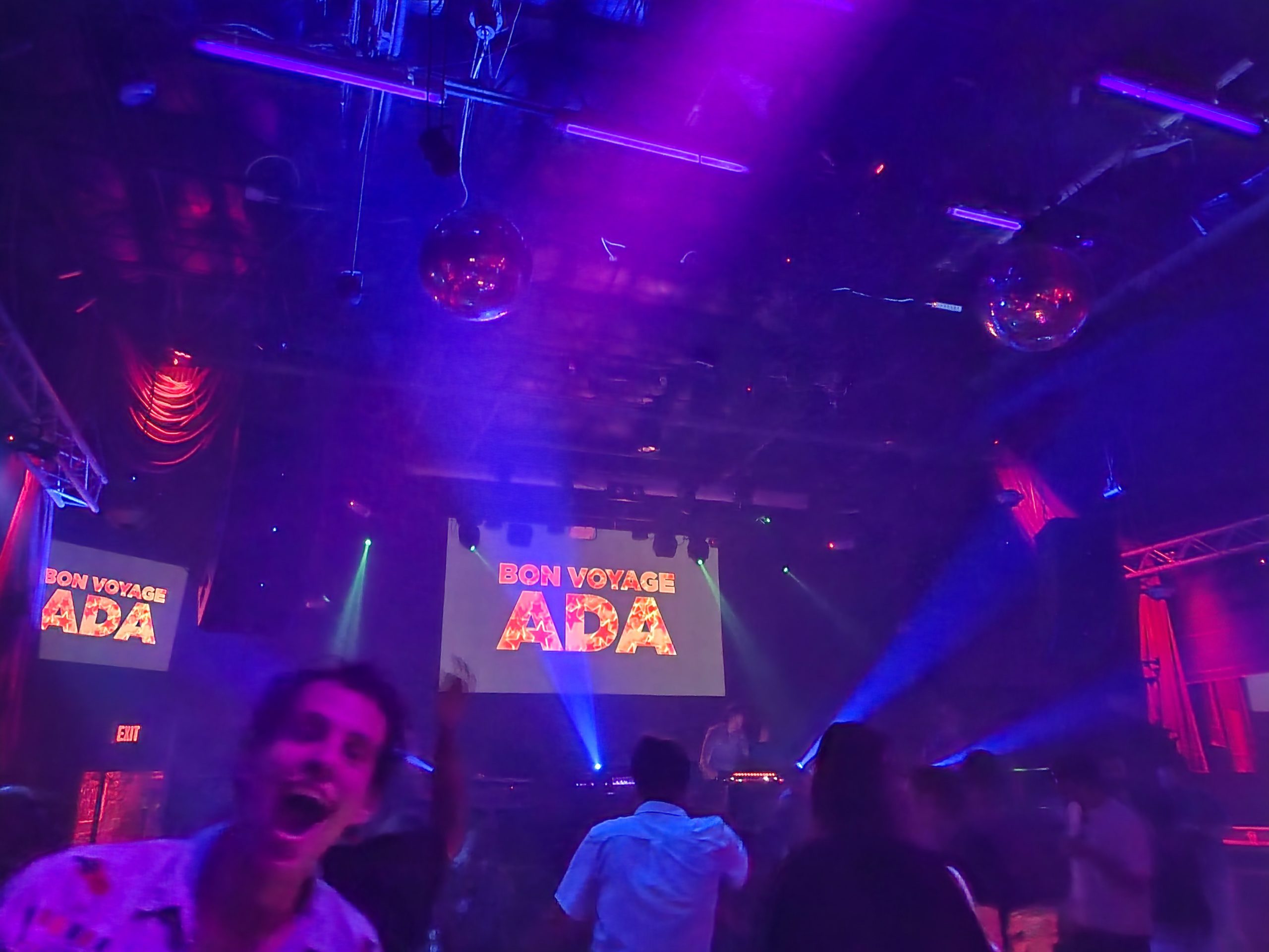 Photo of a dance floor with a message on projected screens "Bon Voyage Ada". An out-of-focus person is looking at the camera with an excited expression.