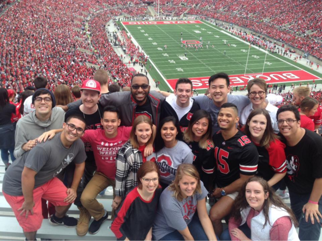 A group of young adults posing in the stands of the Ohio State University football stadium. The field and surrounding stadium seats are visible behind them.