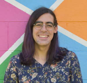 Portrait of Ada in front of their rainbow mosaic garage, April 2021