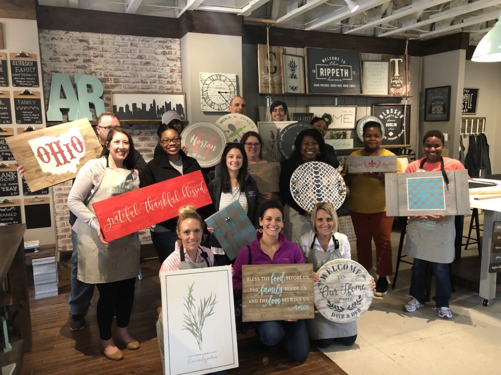 Group of people holding up various wooden wall art, at the AR Workshop in Worthington, OH