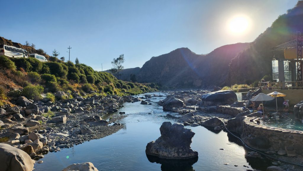 Landscape photograph of the sun setting behind mountains. In the foreground, there is a still and reflective river with rocks. Taken in Colca Canyon, Peru.