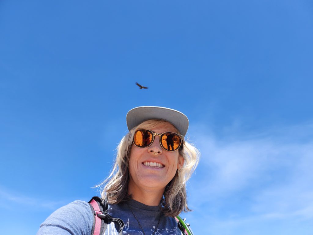 Selfie of Ada with a blue sky background and a condor flying in the distance overhead