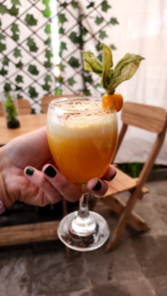 Close-up photo of Ada's hand holding an orange cocktail (Passionfruit Pisco Sour) in a glass, garnished with a goldenberry.