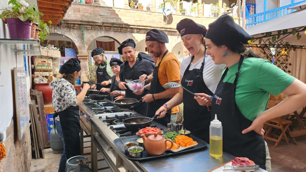 Photo of a group cooking class, with members of Ada's travel group in black aprons and chef's hats, grilling over outdoor stoves in an open courtyard.