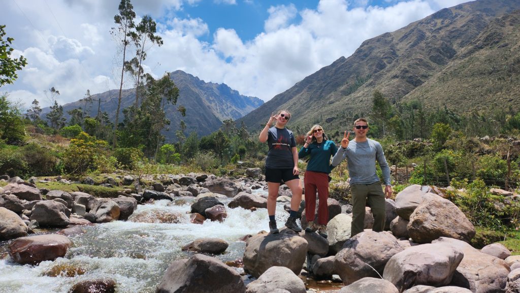 Photo of Ada and their hiking companions on the Lares Trek, standing by a river holding their hands up in the shape of llama heads.