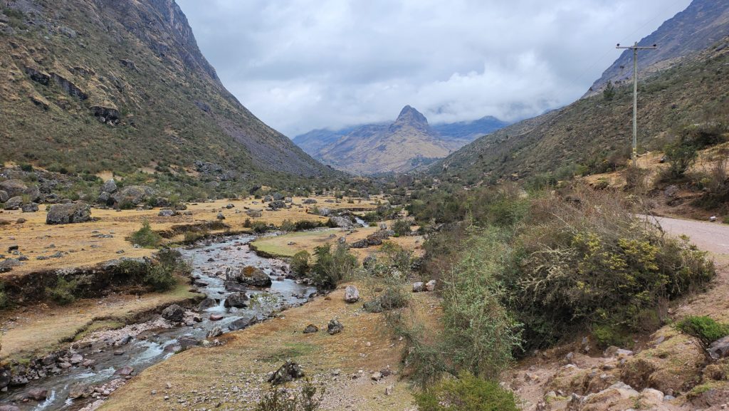 Photo of Urubamba mountain valleys, with cloudy skies, light-green vegetation, and a river winding out toward the distance.