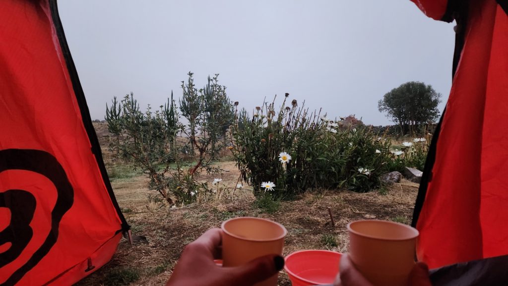 A photo from the inside of a tent, with two hands holding cups of tea in the foreground. The background is a cloudy sky with some small foliage nearby.