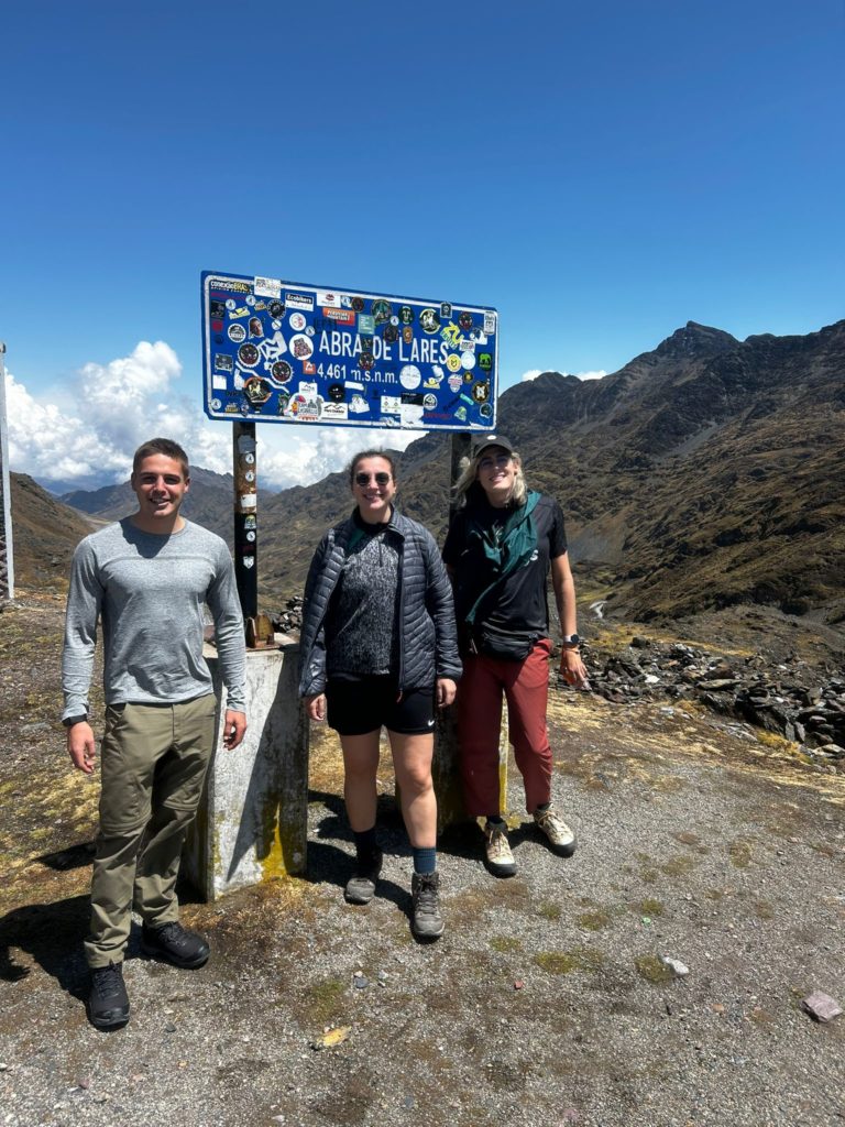 Photo of Ada and their hiking companions on the Lares Trek, standing by a sticker-covered sign in the mountains that reads "Abra de Lares; 4461 m"