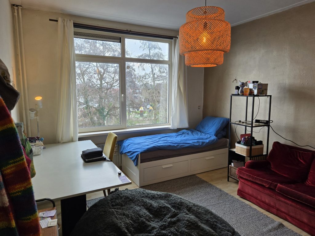 Photo of Ada's bedroom in their first apartment in the Netherlands. There is a large window with white curtains, a red velvet couch, blue-and-white bed, large grey desk, black beanbag chair, and a wood lattice light fixture hanging from the ceiling.