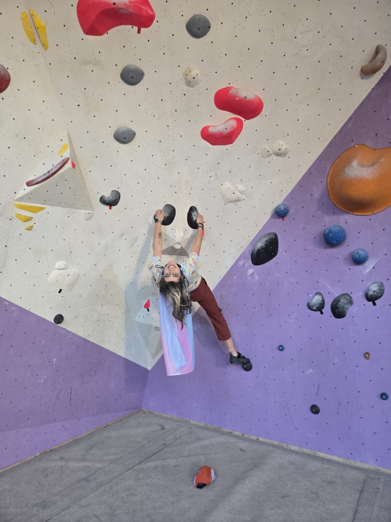 Ada handing upside-down on a climbing wall, wearing a trans flag as a cape.