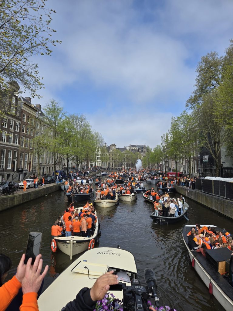 Photo of a canal in amsterdam, full of boats with crowds wearing orange shirts