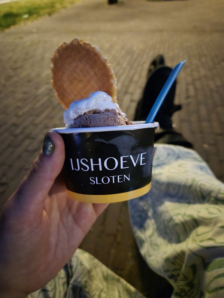 Photo of Ada's hand holding a cup of ice cream from Ijshoeve Sloten. Behind their hand is their leg outstretched, wearing roller blades.
