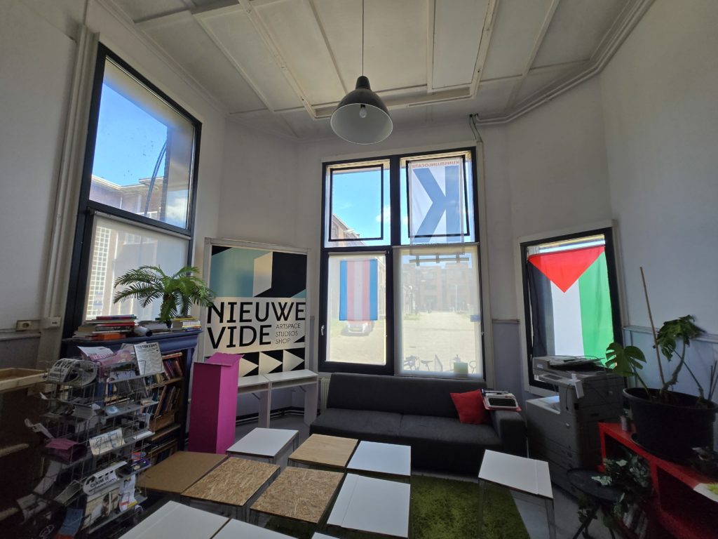 Photo of the interior of the Nieuwe Vide Artspace in Haarlem. There are trans and palestinian flags on the wall, bookshelves, and couches.