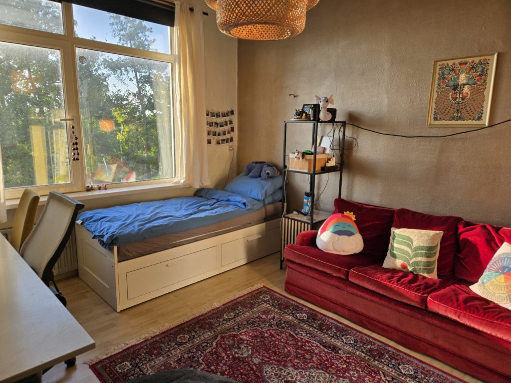 Photo of Ada's bedroom in Haarlem, NL. There is a red velvet couch in front of a bronze-colored wall, their bed, bookshelf, and desk. There is a window with a view of trees, art on the walls, and a brown lattice light fixture overhead.