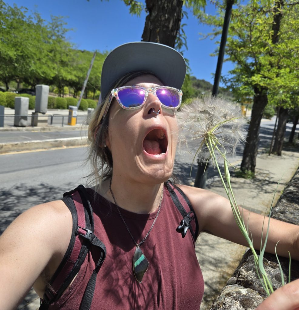 Photo of Ada, wearing sunglasses, a tanktop, and backpack, pretending to eat a dandelion the size of their head