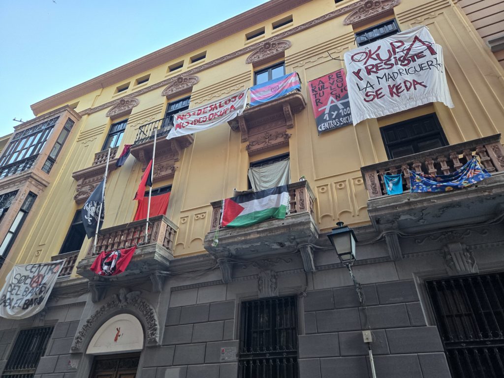 Photo of a squatted building in Spain, which has balconies adorned with various pride and protest flags.