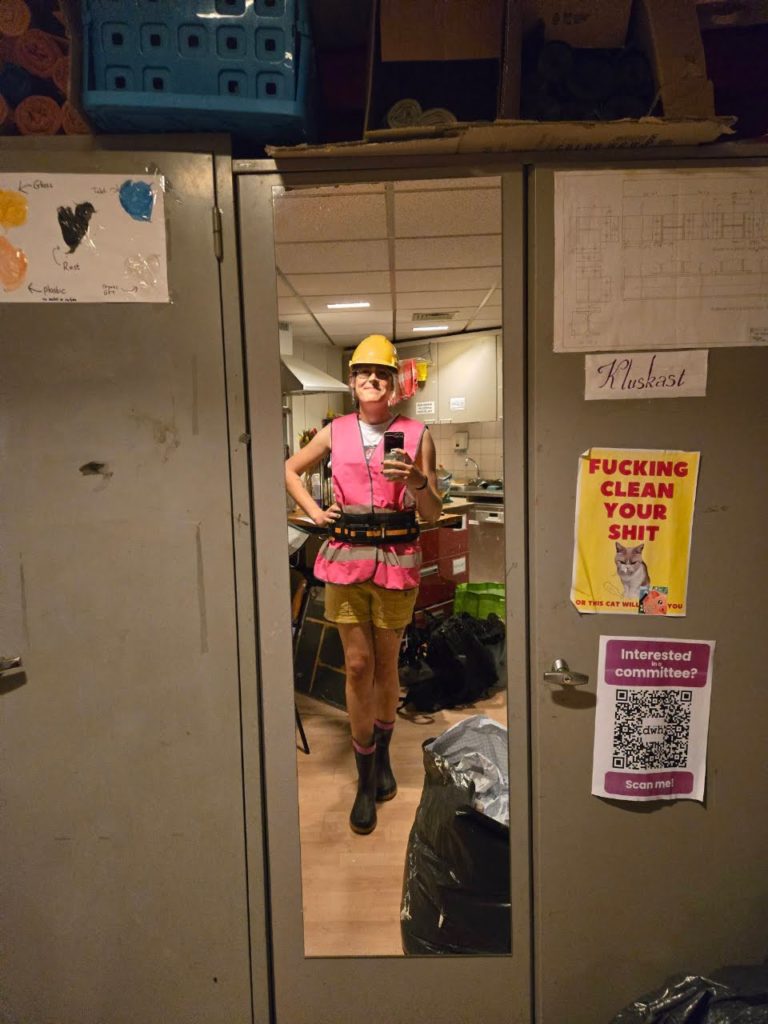 Photo of Ada in front of a mirror, wearing a cute construction-themed outfit for their drag performance