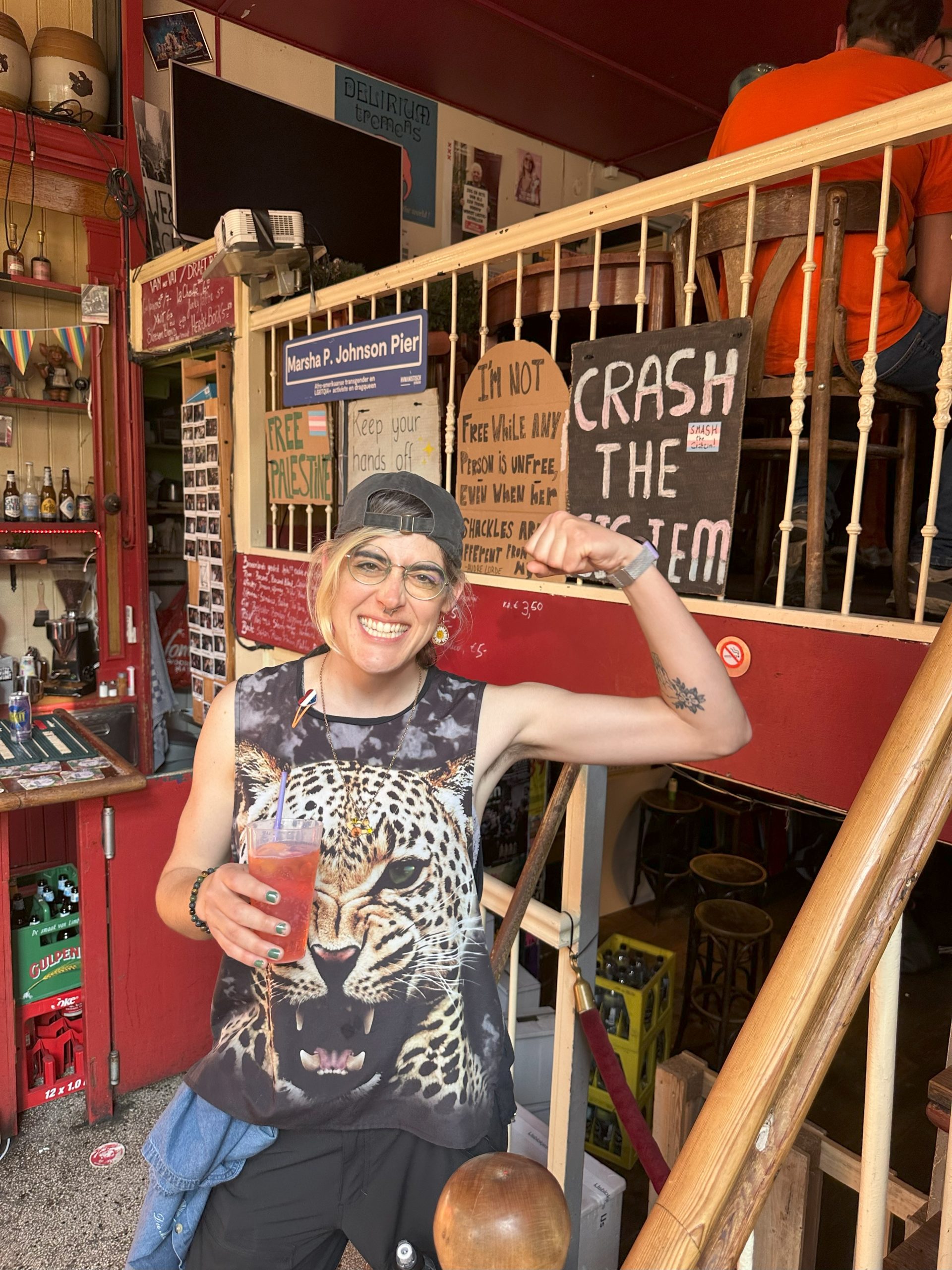 Photo of Ada inside Cafe Saarein in Amsterdam, flexing and holding a drink in front of a sign that says "Crash the CIStem"