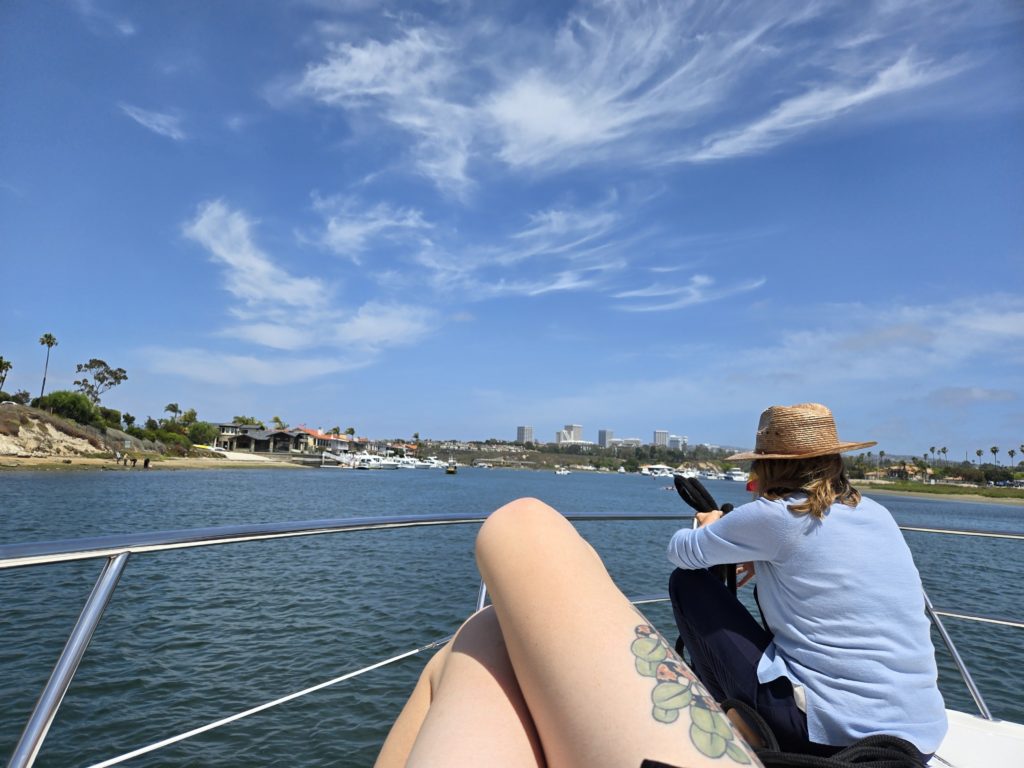 Photo of the view from the bow of a small yacht, inside Newport Harbor. Ada's legs and aunt are in the foreground.