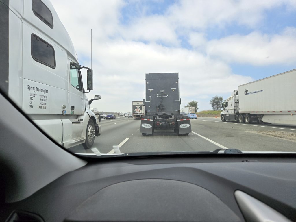 Picture of the view from a car windshield. There are semi trucks in front of, to the left, and to the right of our view.